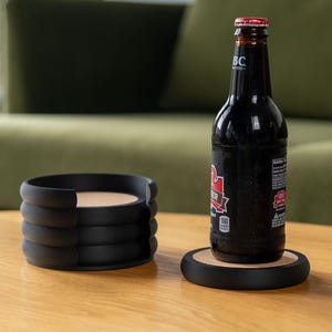 Op de afbeelding: Een donkere bierfles met een rode dop staat op een zwarte onderzetter met een kurken midden. Daarnaast een stapel zwarte onderzetters met een vergelijkbaar ontwerp. De onderzetters staan op een lichtbruine houten tafel.