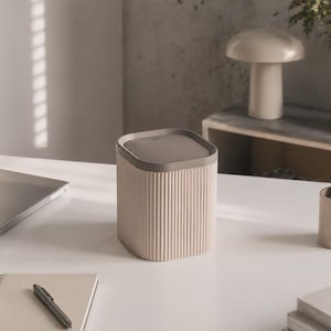 Puede incluir: Una papelera pequeña de forma cuadrada con un exterior de color beige claro acanalado y una tapa plana de color gris oscuro. El recipiente presenta ranuras verticales en los lados y un borde superior liso y redondeado, con un diseño funcional.
