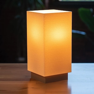 Pode incluir: Um candeeiro de mesa retangular com um abajur de tecido texturizado em branco sujo que emite um brilho quente e suave. O candeeiro é suportado por uma base quadrada sólida que proporciona uma fundação estável à estrutura vertical e quadrada.