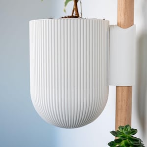 May include: A white cylindrical planter with a vertical ribbed texture attached to a light-coloured wooden pole. The planter holds a small green plant with variegated leaves, and a separate green succulent is visible below the planter on the pole.