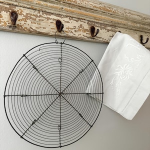 May include: A black, circular wire cooling rack hangs from a distressed, off-white wooden rack with metal hooks. A white, embroidered linen towel is to the right of the rack. The wall is painted a light gray.