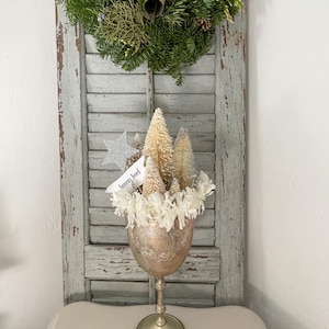 May include: A festive Christmas arrangement in a gold-toned goblet. The goblet holds miniature bottle brush trees, white paper flowers, and a silver star. A small tag reads "Joyeux Noel". A green wreath with a bell hangs in the background.