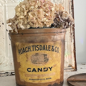 May include: A vintage brown metal candy container with a yellow label that reads "Roach, Tisdale & Co. Candy Minneapolis, Minn.". The container is decorated with a black and white image of a building. Dried flowers are in the container.