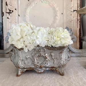 May include: A rectangular, distressed, silver-colored planter filled with white hydrangea flowers. The planter has ornate detailing and sits on small feet. The background includes a distressed white door and decorative elements.