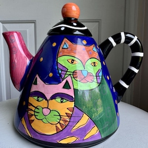May include: A colorful ceramic teapot with a whimsical cat design. The teapot features a blue background with orange, green, and purple cat faces. The handle is black with white stripes, and the spout is pink and red.