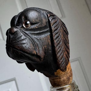 May include: A hand-carved wooden bottle stopper featuring a dog's head. The dog's head is dark brown with a detailed carved texture, and a clear, round eye. The stopper is attached to a glass bottle with a silver-colored metal band.