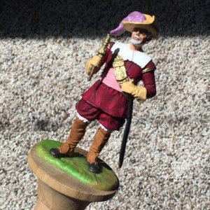 May include: A plastic toy figurine of a man wearing a red and white costume with a purple hat and holding a sword. The figurine is standing on a wooden base with green grass.
