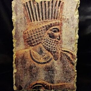 May include: A close-up of a stone relief sculpture of a man with a crown and beard. The sculpture is carved in a reddish-brown stone and has a textured surface.