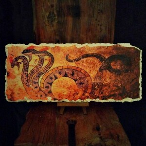 May include: A rectangular artwork featuring a stylized snake with three heads and a coiled body. The artwork is rendered in shades of orange, brown, and purple on a textured, off-white background. The piece is displayed on a small wooden easel.