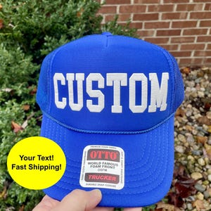 May include: A blue trucker hat with the word "CUSTOM" in white block letters. The hat has a mesh back and a curved brim. The hat also has a tag that says "OTTO" and "TRUCKER".