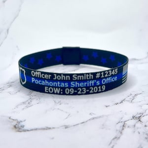 May include: Black and blue elastic bracelet with a thin blue line, stars, and the text "Officer John Smith #12345 Pocahontas Sheriff's Office EOW: 09-23-2019".