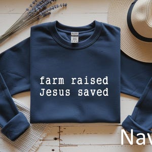 May include: Navy blue sweatshirt with the text "farm raised Jesus saved" in white. A straw hat and lavender sprigs are in the background. The word "Navy" is also visible.