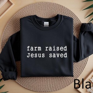 May include: Black crewneck sweatshirt with the text "farm raised Jesus saved" in white. The sweatshirt is displayed on a woven mat with a wooden tray and greenery in the background. The word "Black" is visible.