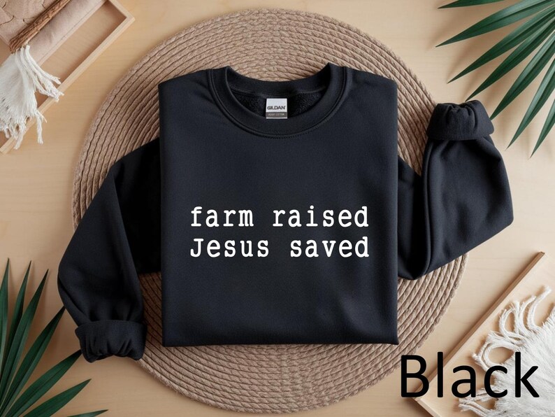 May include: Black crewneck sweatshirt with the text "farm raised Jesus saved" in white. The sweatshirt is displayed on a woven mat with a wooden tray and greenery in the background. The word "Black" is visible.