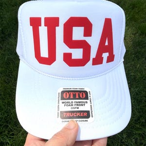 May include: White trucker hat with the red letters "USA" across the front. The hat has a mesh back and a curved brim. The hat has a "OTTO" label on the front.