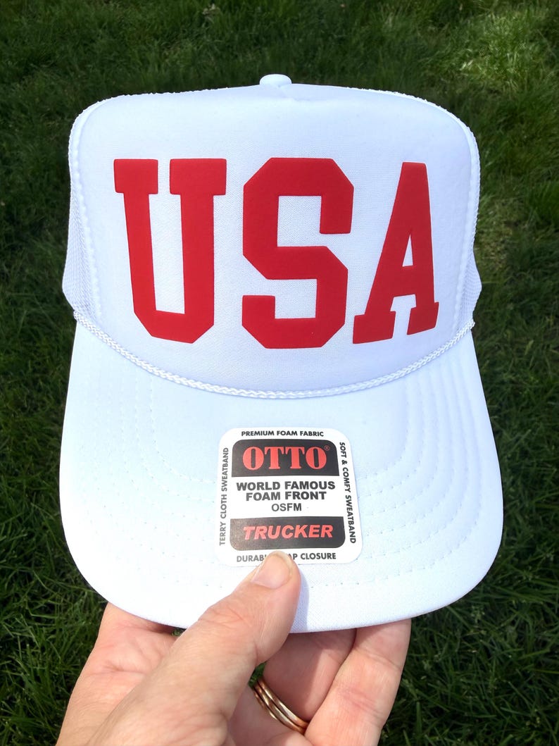 May include: White trucker hat with the red letters "USA" across the front. The hat has a mesh back and a curved brim. The hat has a "OTTO" label on the front.