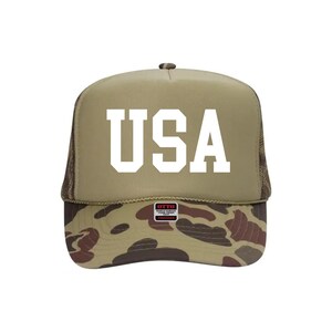 May include: A tan trucker hat with the word "USA" in bold white letters. The brim features a camouflage pattern in shades of brown, green, and tan. The hat has a mesh back.