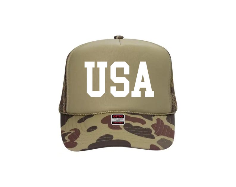 May include: A tan trucker hat with the word "USA" in bold white letters. The brim features a camouflage pattern in shades of brown, green, and tan. The hat has a mesh back.