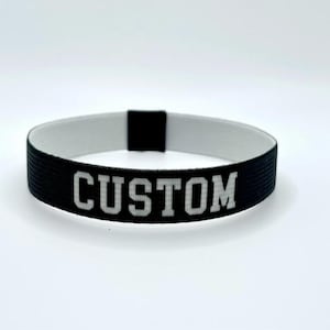Op de afbeelding: Zwart-witte elastische polsband met het woord "CUSTOM" in witte, vette letters. De polsband is aan de buitenkant zwart en aan de binnenkant wit. De tekst staat in het midden van de band.