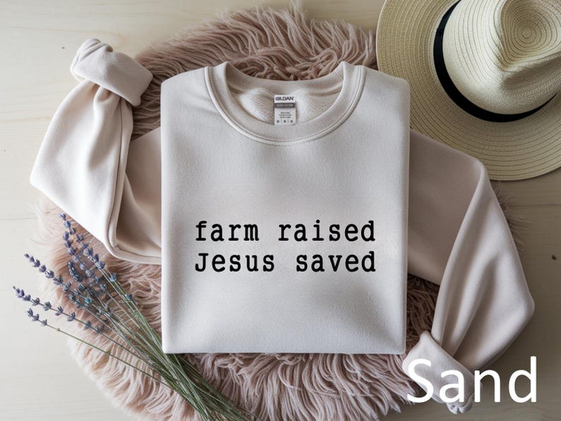 May include: Sand-colored sweatshirt with the text "farm raised Jesus saved" in black. The sweatshirt is folded on a fluffy, light brown surface, with a straw hat and a bundle of lavender in the background.