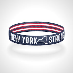 Op de afbeelding: Een blauw en wit armband met rode strepen. Het armband zegt "NEW YORK STRONG" met een omtrek van de staat New York.