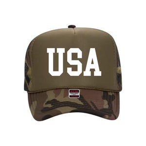 May include: A trucker hat with a camouflage brim and a khaki-colored crown. The word "USA" is printed in bold white letters on the front. The hat has a mesh back and a small tag on the brim.