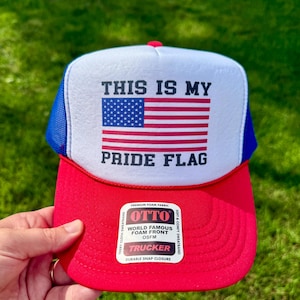 Op de afbeelding: Een rode, witte en blauwe truckerpet met de tekst "THIS IS MY PRIDE FLAG" en een Amerikaanse vlagafbeelding. De pet heeft een witte schuim voorkant, een rode rand en een blauwe mesh achterkant. De pet wordt door een hand vastgehouden.