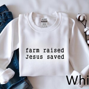 May include: A white crewneck sweatshirt with the text "farm raised Jesus saved" in black. The word "White" is written in black at the bottom right. Blue jeans are visible in the background.
