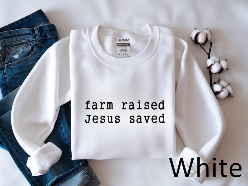 May include: A white crewneck sweatshirt with the text "farm raised Jesus saved" in black. The word "White" is written in black at the bottom right. Blue jeans are visible in the background.