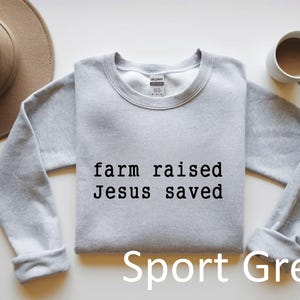 May include: A light grey sweatshirt with the text "farm raised Jesus saved" in black. The words "Sport Grey" are printed below the text. A brown hat and a white mug with a brown liquid are also in the image.