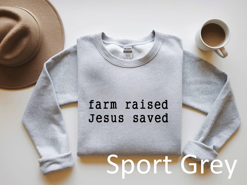 May include: A light grey sweatshirt with the text "farm raised Jesus saved" in black. The words "Sport Grey" are printed below the text. A brown hat and a white mug with a brown liquid are also in the image.