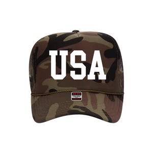 May include: Camouflage trucker hat with the word "USA" in bold white letters. The hat features a mesh back and a curved brim. The hat has a small tag that says "OTTO".
