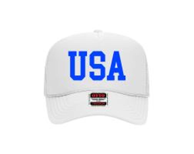 May include: White trucker hat with the word "USA" in blue block letters across the front. The hat has a structured front panel and a mesh back. A small tag is visible on the brim.