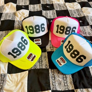 以下が含まれることがあります： 黒い文字で「1986」とプリントされた白いフォームフロントパネルを持つ4つのトラッカーキャップ。メッシュの背面と、ネオンイエロー、ブラック、ホットピンク、ブライトブルーのつばが特徴です。各キャップのつばにはOttoブランドのステッカーが貼られています。