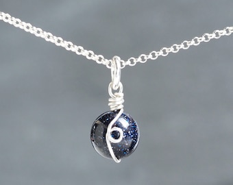 May include: A silver chain necklace featuring a dark blue, spherical pendant. The pendant is wire-wrapped and has a speckled, glittery appearance. The necklace is set against a grey backdrop.
