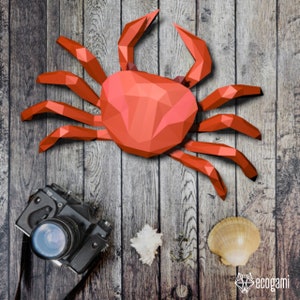 May include: A red origami crab on a wooden background. The crab is made of paper and has a detailed design. There is a camera and a seashell in the lower right corner of the image.