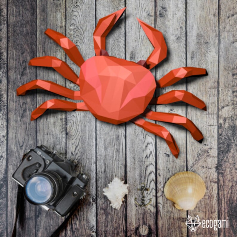 May include: A red origami crab on a wooden background. The crab is made of paper and has a detailed design. There is a camera and a seashell in the lower right corner of the image.