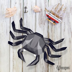 May include: A black papercraft crab with a detailed, geometric design. The crab is posed with its legs outstretched, as if it is walking. The crab is on a white wooden background.