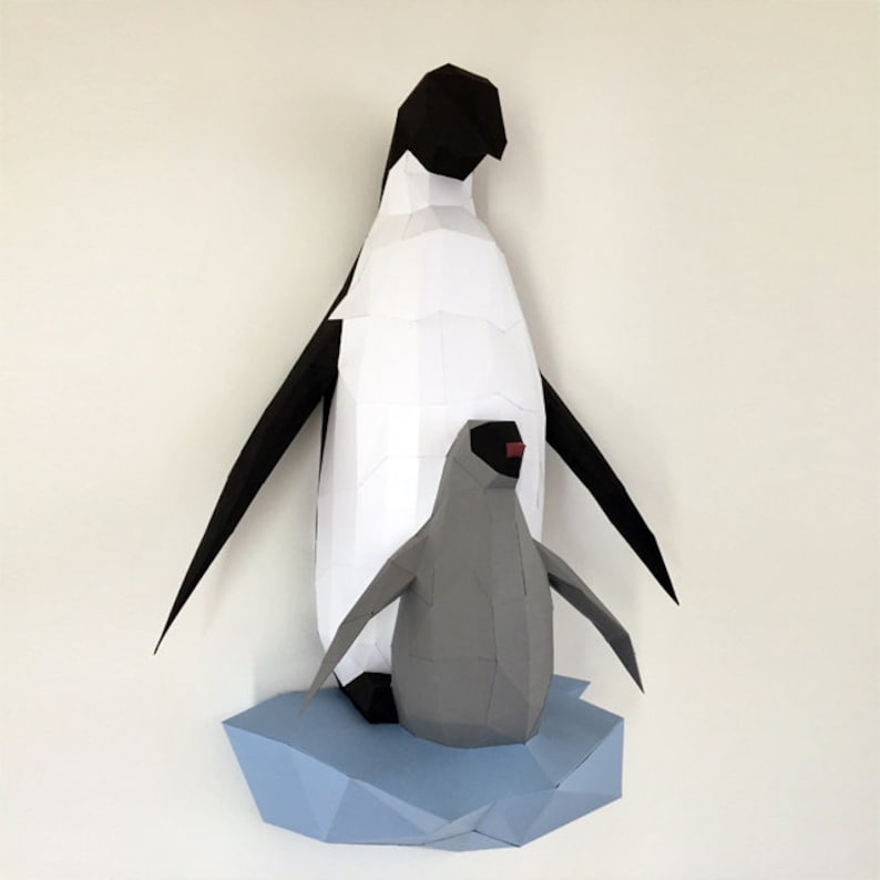 May include: A papercraft model of two penguins standing on a blue paper ice floe. The larger penguin is black and white, while the smaller penguin is gray. Both penguins have red beaks.