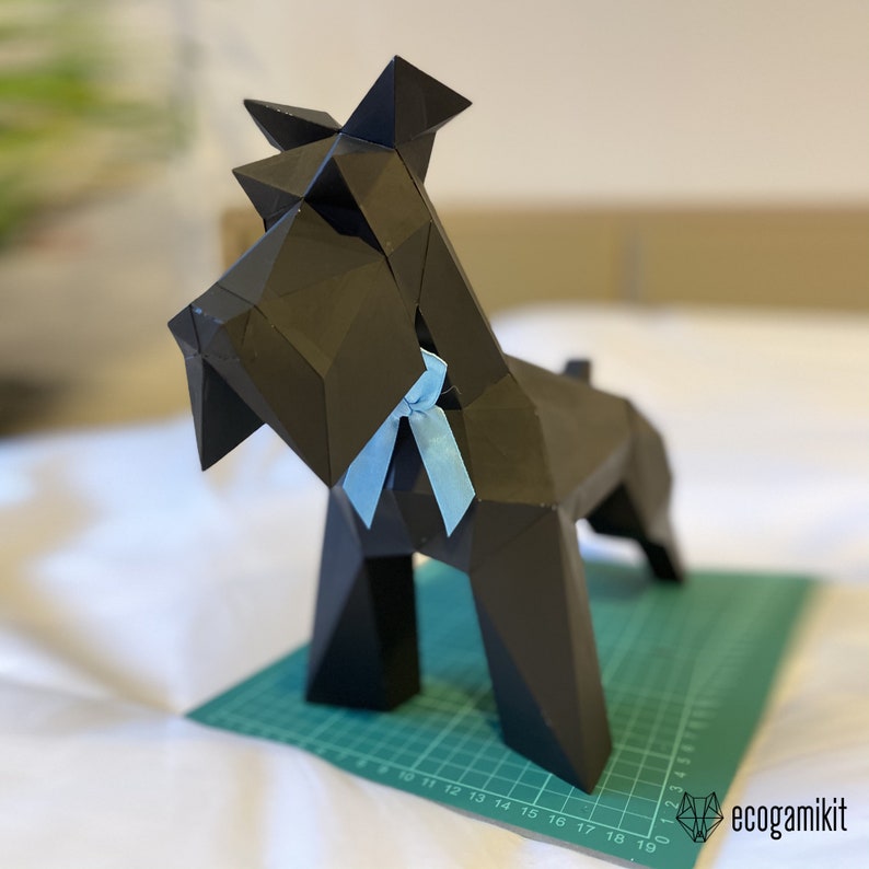 K&ouml;nnte beinhalten: Ein schwarzes Papiermodell eines Schnauzer-Hundes mit einer hellblauen Schleife. Das Modell steht auf einer gr&uuml;nen Schneidematte mit Markierungen in Zentimetern.