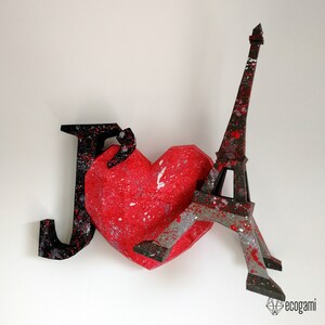 May include: A black and red paper mache heart with a black paper mache Eiffel Tower leaning against it. The heart and tower are decorated with black and white paint splatters.
