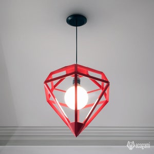 May include: Red geometric pendant light fixture with a diamond-like shape. The light has a black ceiling mount and a single exposed light bulb. The fixture is suspended from the ceiling by a black cord. The light is turned on.