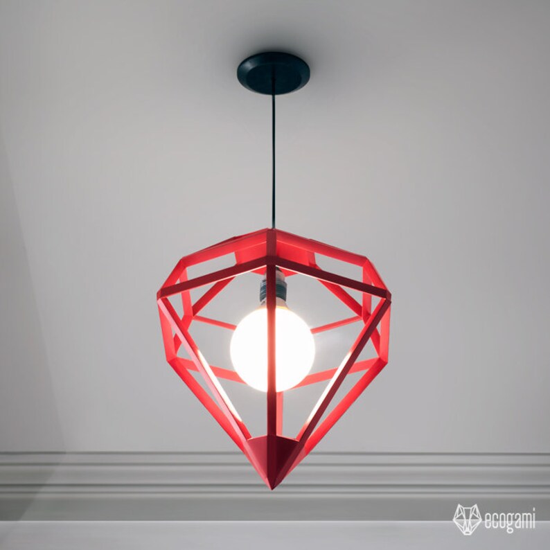 May include: Red geometric pendant light fixture with a diamond-like shape. The light has a black ceiling mount and a single exposed light bulb. The fixture is suspended from the ceiling by a black cord. The light is turned on.