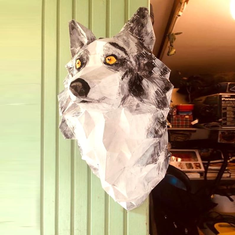 Peut inclure: Une sculpture de t&ecirc;te de loup blanche et grise avec des yeux jaunes. La sculpture est en papier et est peinte dans un style r&eacute;aliste. Elle est mont&eacute;e sur un mur.