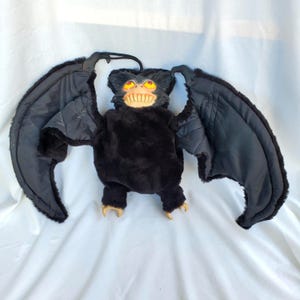May include: A plush bat toy featuring black faux fur, large outstretched wings with black fabric undersides, and a molded plastic face with yellow eyes and prominent white teeth. The creature has small claws on its feet and a hanging loop.