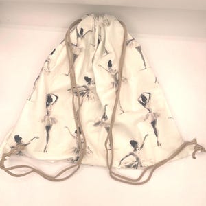 May include: A drawstring backpack made of off-white fabric featuring a repeating black and white print of ballet dancers in various poses. The bag includes two tan cord straps that cinch the top closed when pulled tight.