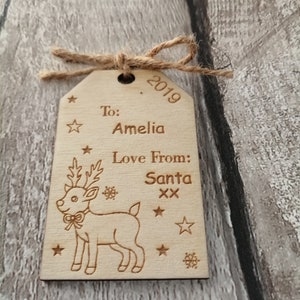 May include: A wooden Christmas gift tag with a reindeer illustration. The tag is personalized with the text "To: Amelia Love From: Santa xx" and the year "2019".