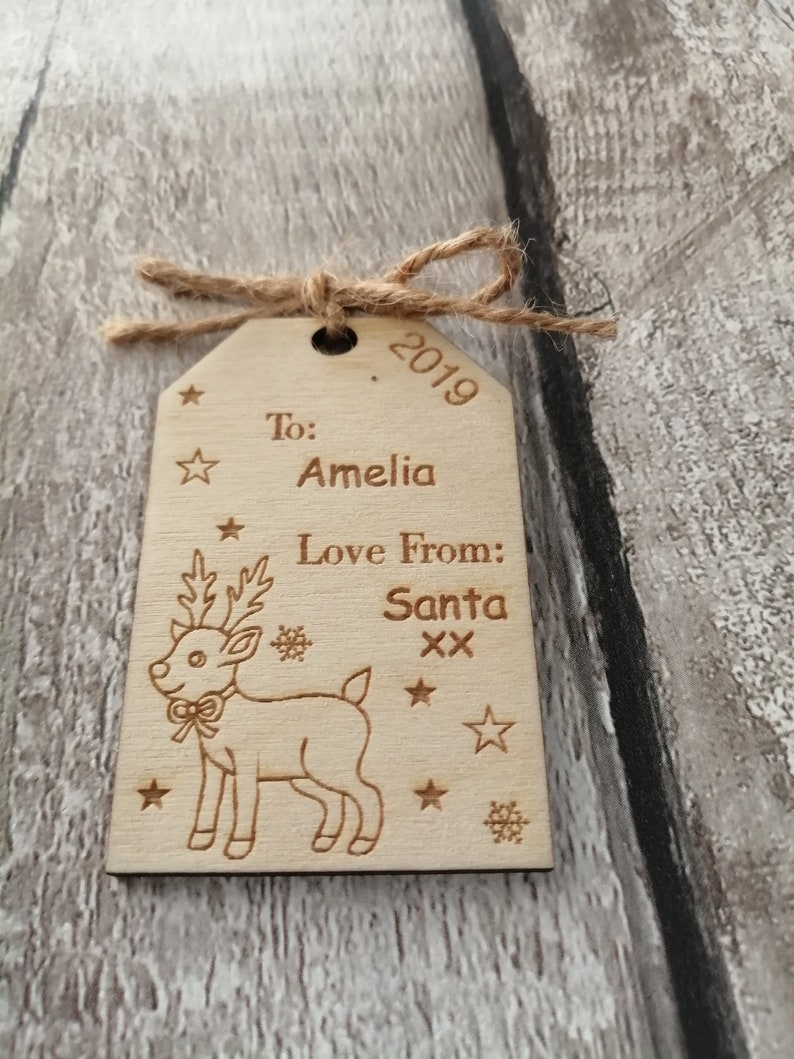 May include: A wooden Christmas gift tag with a reindeer illustration. The tag is personalized with the text "To: Amelia Love From: Santa xx" and the year "2019".