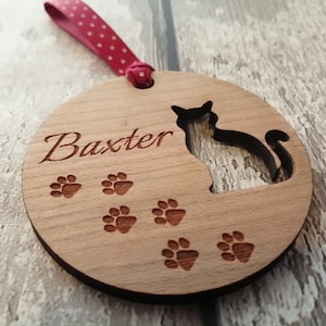May include: A round wooden bauble with a cat silhouette, paw prints, and the name "Baxter" engraved on it. The bauble is attached to a red and white polka dot ribbon.