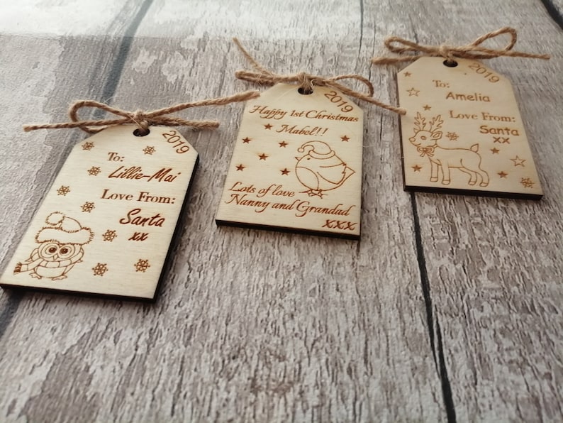 May include: Three wooden Christmas gift tags with twine. The tags are decorated with a reindeer, an owl, and a bird. The tags are personalized with the names Amelia, Lillie-Mai, and Mabel. The tags also include the year 2019 and messages of love from Santa.
